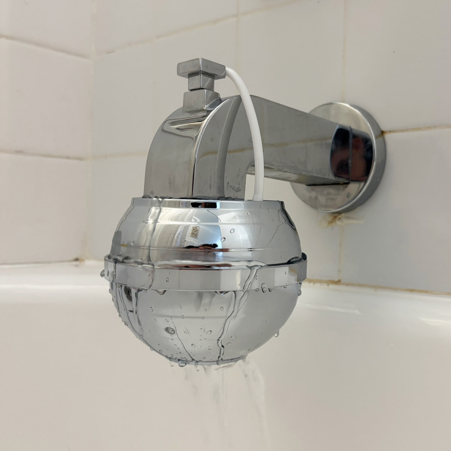 A chrome Tru Bath Water Filter hanging from a chrome bath spout against a white tile background. (Courtesy Zoe Malin)