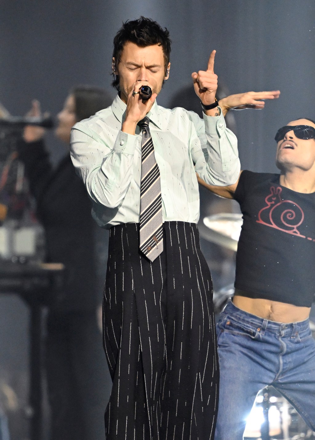 Harry Styles performing on stage, dressed in a light-colored shirt and striped tie, holding a microphone.