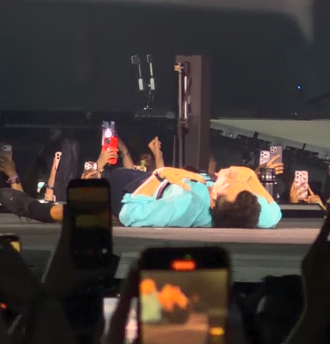 Harry Styles lies on a stage in a blue shirt with his hands covering his face, surrounded by people holding up phones.