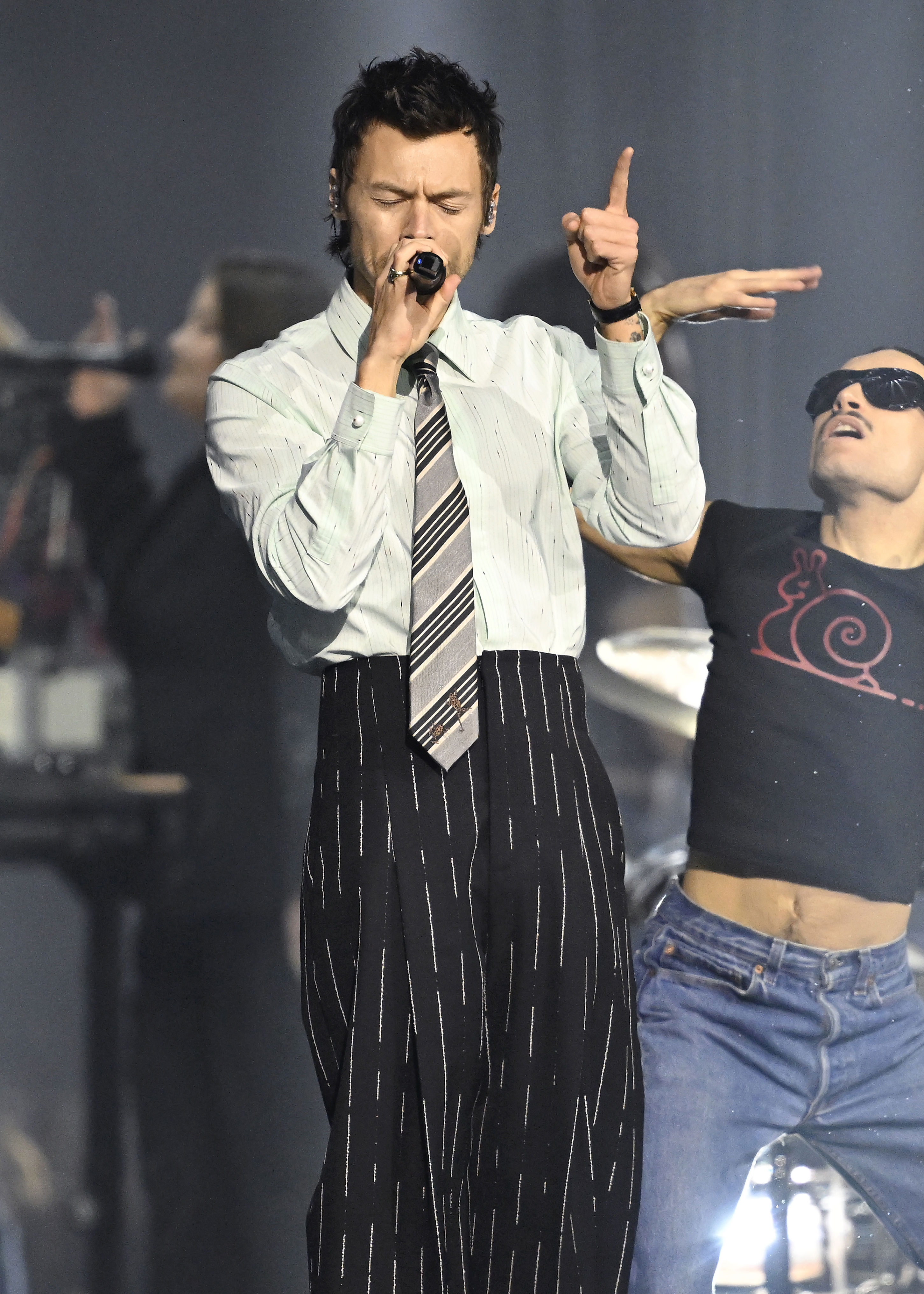 Harry Styles performing on stage, dressed in a light-colored shirt and striped tie, holding a microphone.