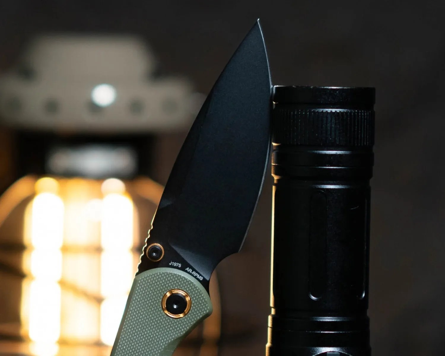 Black folding knife with green textured handle leaning against a black flashlight with bright light in the background.