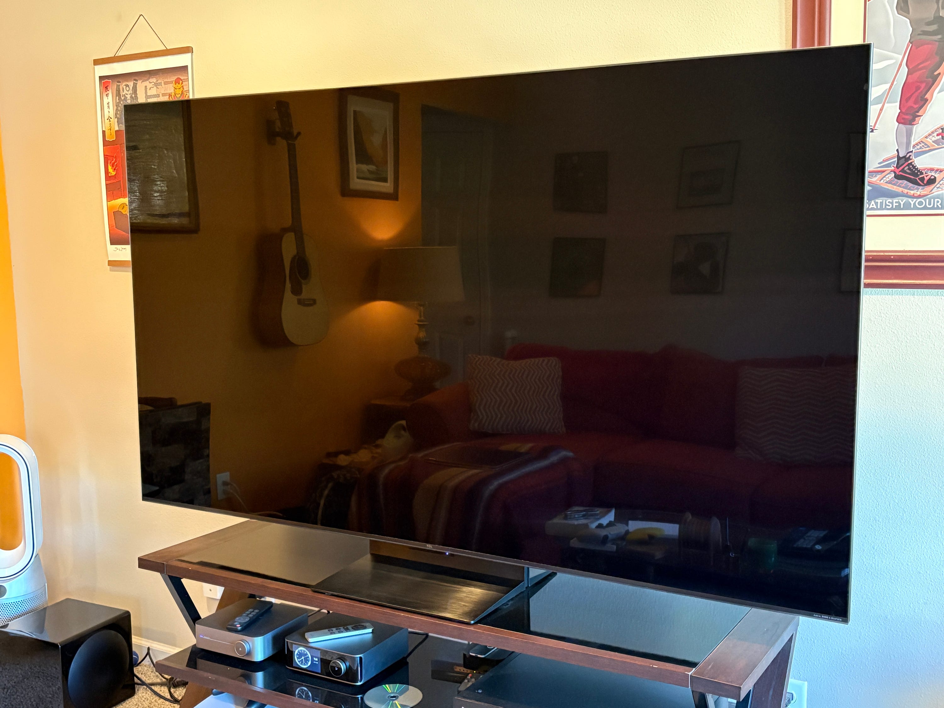 A TCL QM7L on a media stand reflects a living room with a couch, a lamp, wall art, and a guitar.