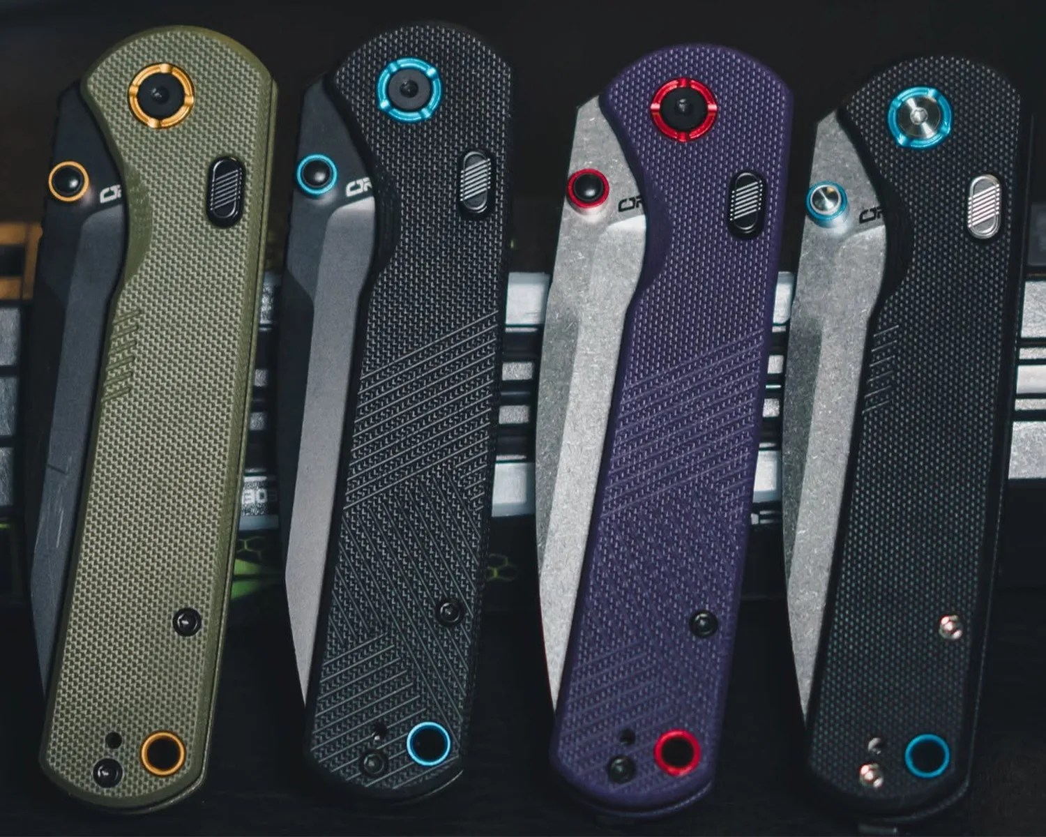 Four folding knives with textured handles in olive green, black, purple, and black, each with colored accents on screws and buttons.