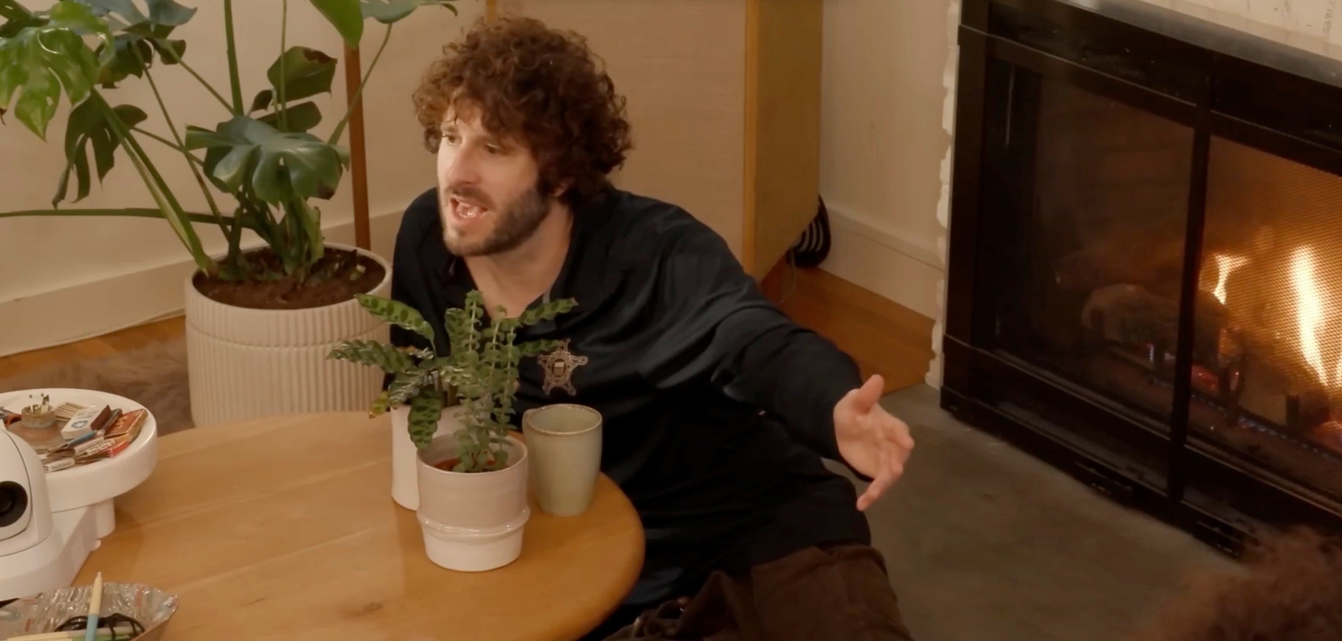 Man sitting at a dining table with plants, gesturing while talking, near a lit fireplace indoors