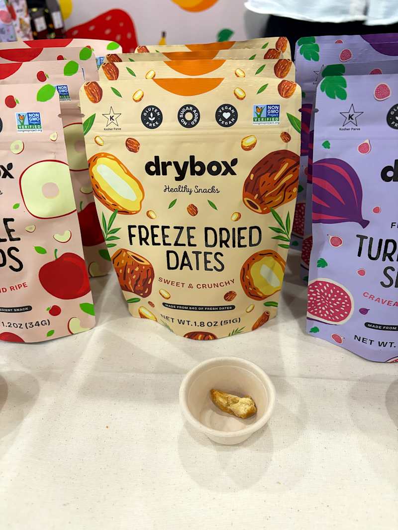 Pouches of dry snacks including freeze-dried dates, with a sample piece in a small cup on a table.