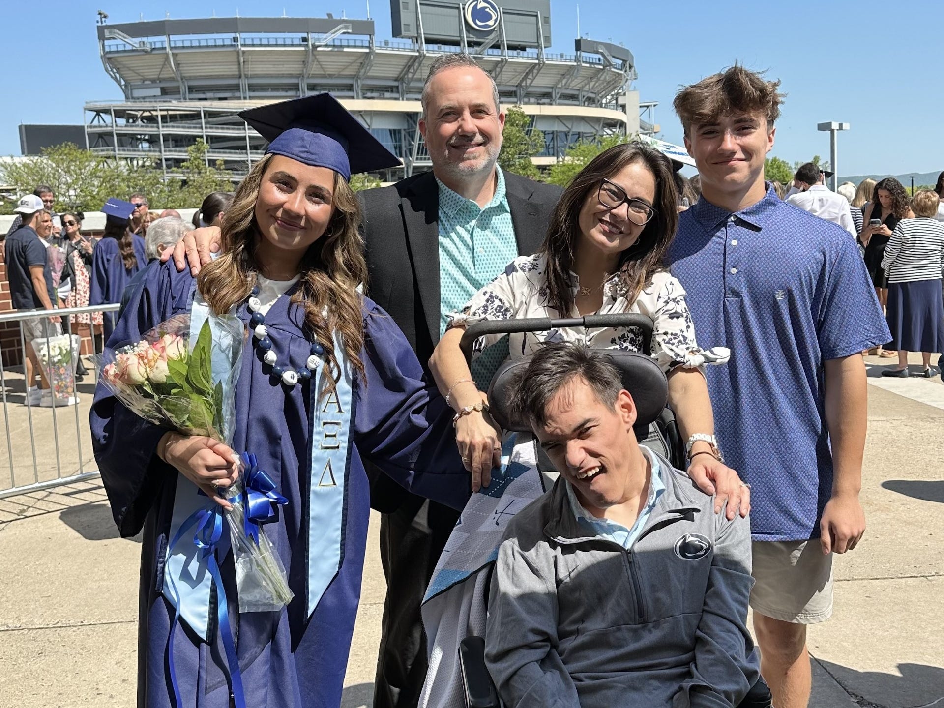 Group poses with a graduate in blue cap and gown outside a Penn State stadium on a sunny day.