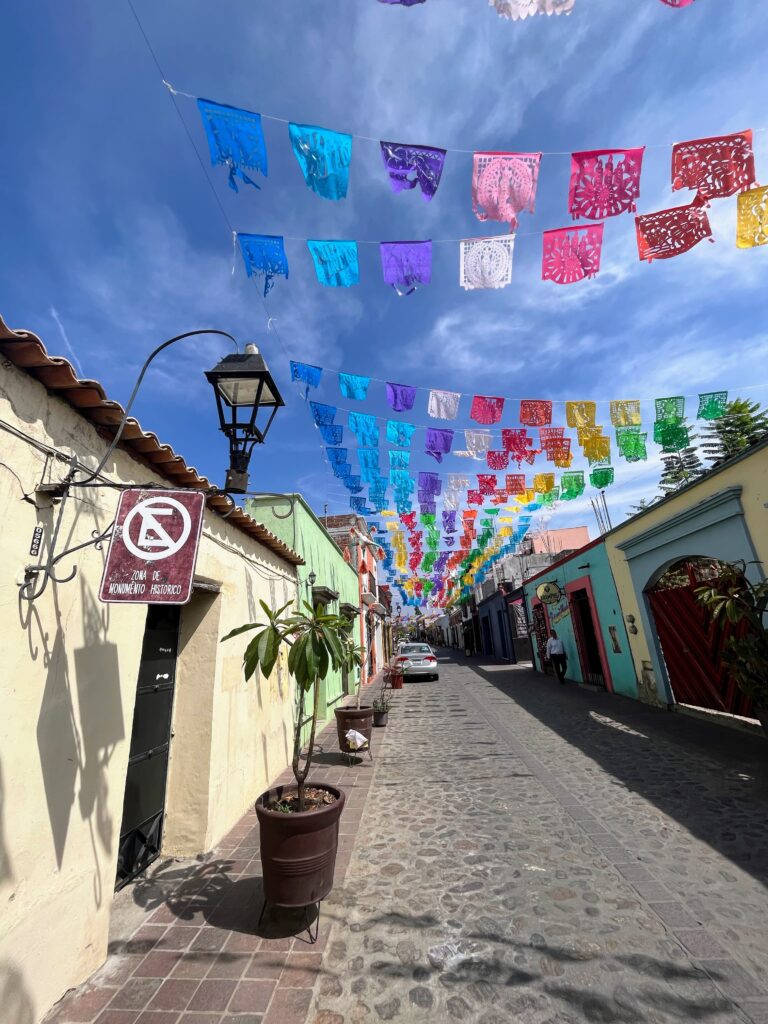 Rows of vibrant papel picado banners in blue, purple, pink, red, yellow, and green stretch overhead across a narrow cobblestone street lined with pastel buildings. A vintage streetlamp and potted plants add charm beneath a dramatic blue sky streaked with wispy clouds.