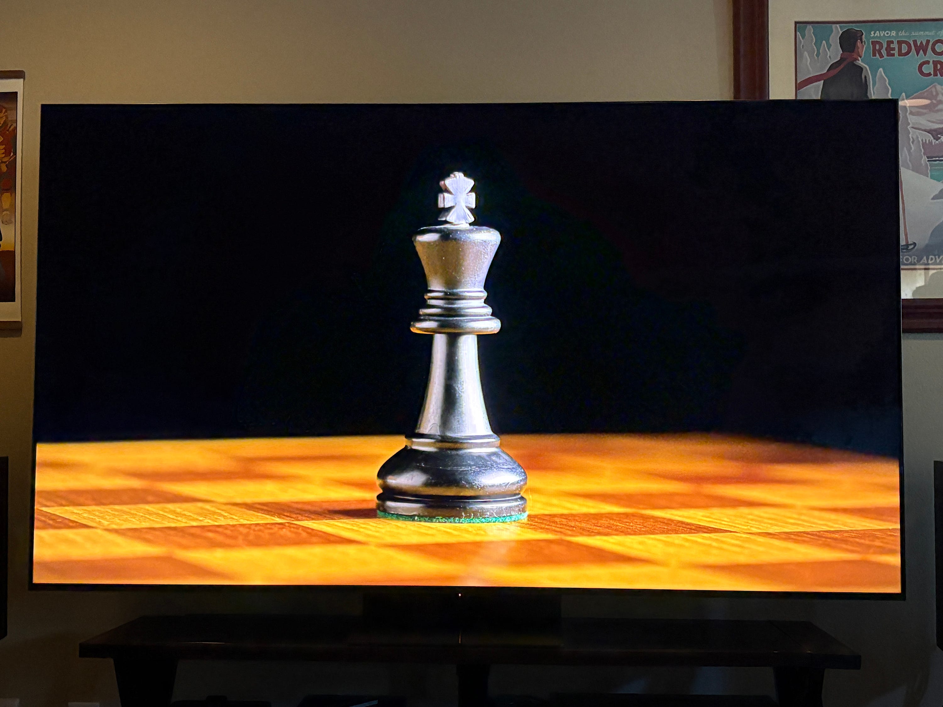 A TCl QM7L TV displays a close-up of a black chess king on a wooden chessboard against a dark background.