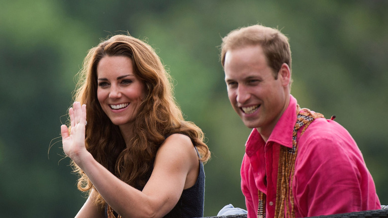 Kate Middleton wearing a blue top waving next to Prince William wearing a red shirt.