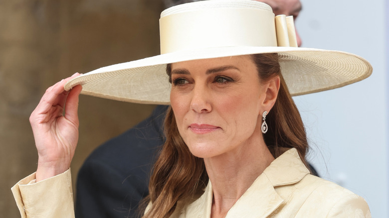 Kate Middleton touching the brim of her yellow hat wearing a matching blazer with diamond earrings.