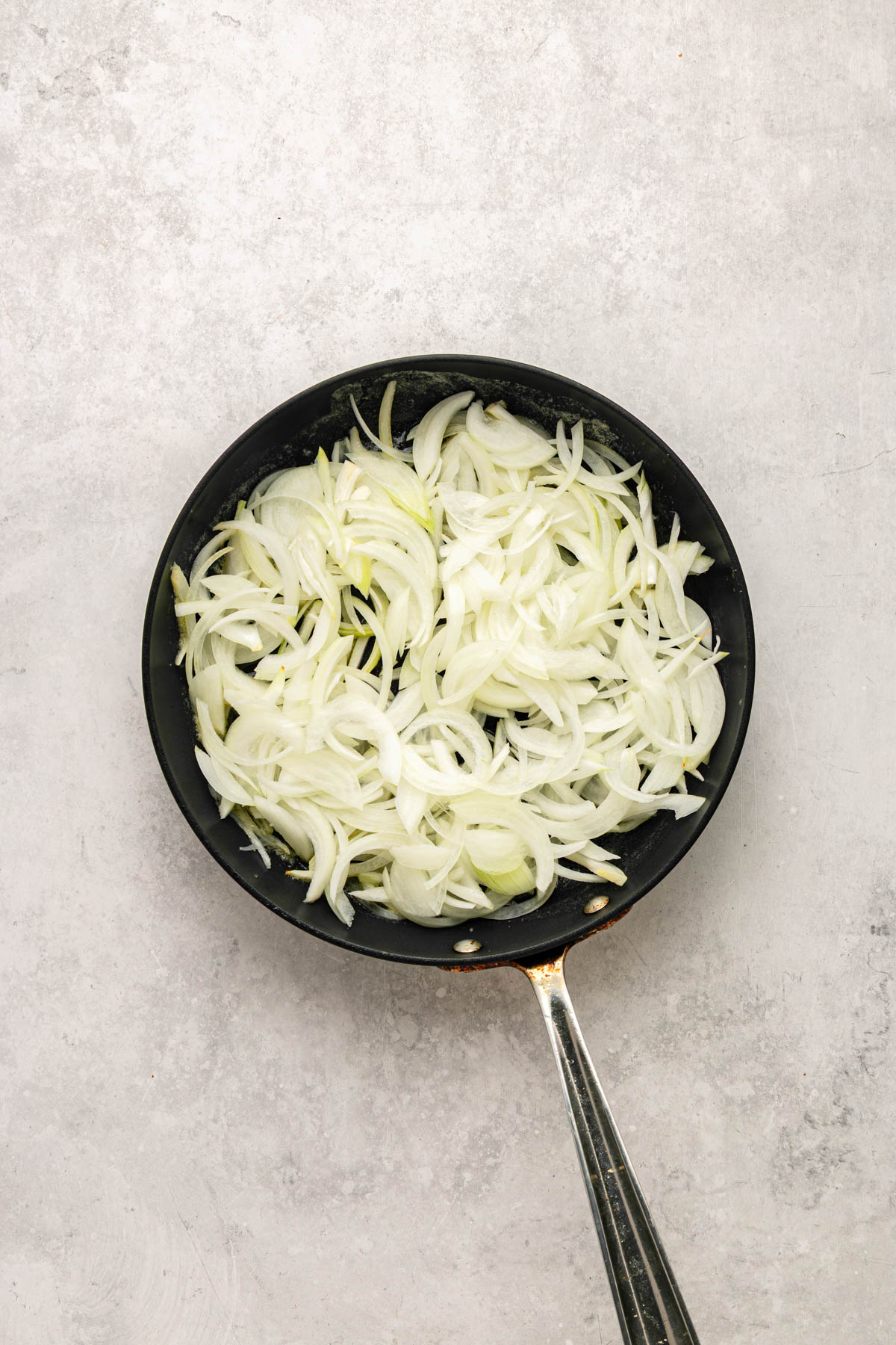 A black frying pan filled with sliced raw onions is placed on a light gray textured surface.