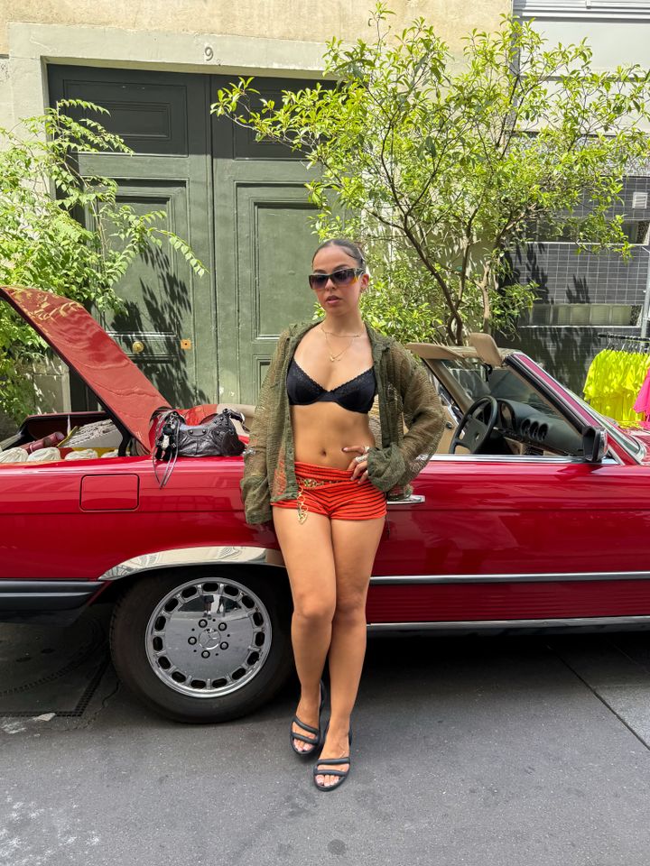 A person posing in front of a convertible car with an open hood on a city street
