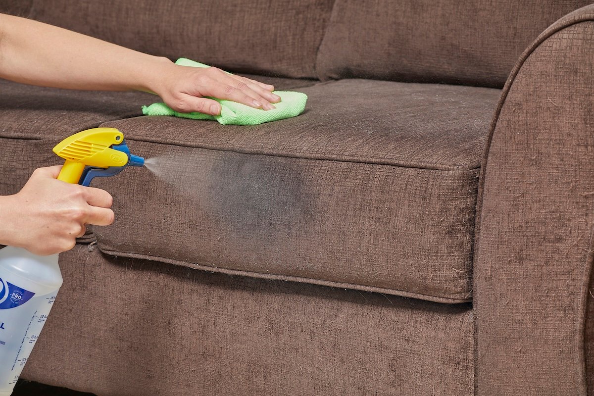 Woman cleans a brown couch with a spray bottle in one hand and a microfiber cloth in another.