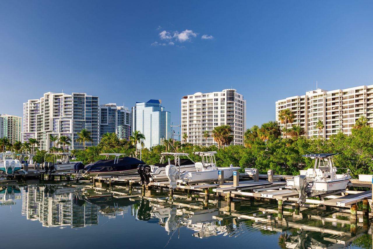 A marina with boats docked in front of a backdrop of modern highrise buildings and greenery