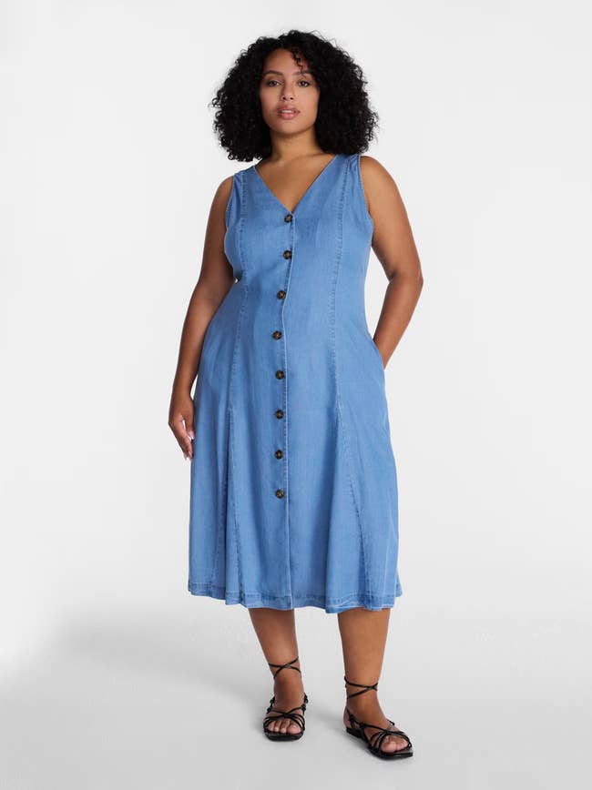 Person wearing a sleeveless denim button-up dress with sandals, standing against a plain background