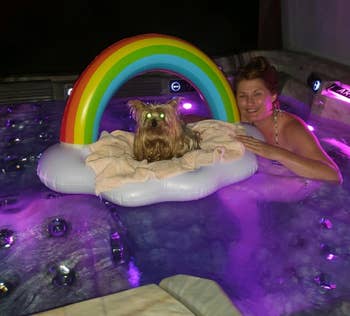 a person in a hot tub with a small dog on a rainbow cloud float, surrounded by colorful lights