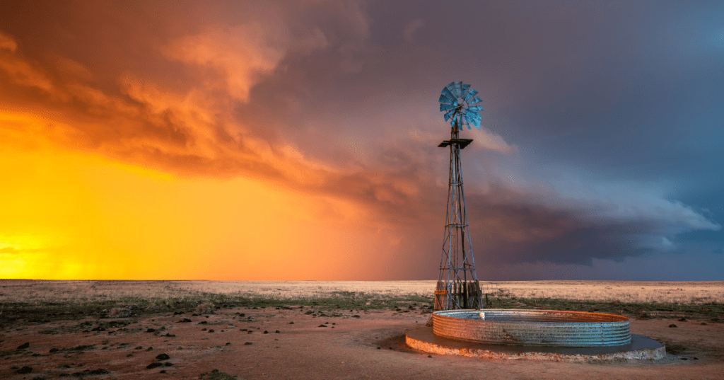 a windmill sits in the middle of a field with a sunset and thunderstorm looming overhead in rural kansas