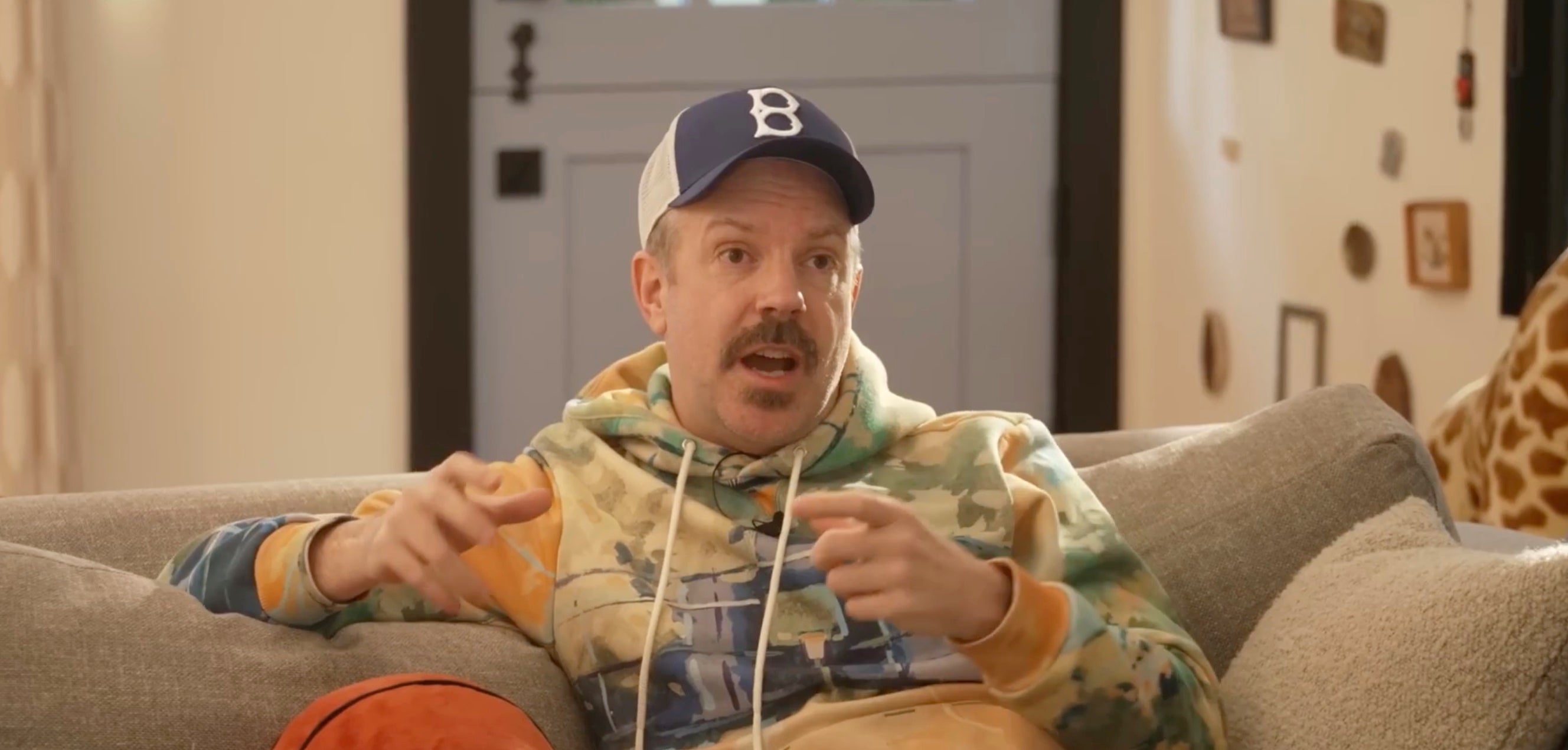 Person wearing a baseball cap and graphic hoodie sits on a couch, gesturing while speaking