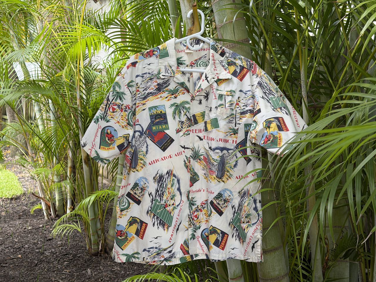 A vintage short-sleeve shirt with printed designs of Florida themed imagery hanging on a hanger amidst tropical plants