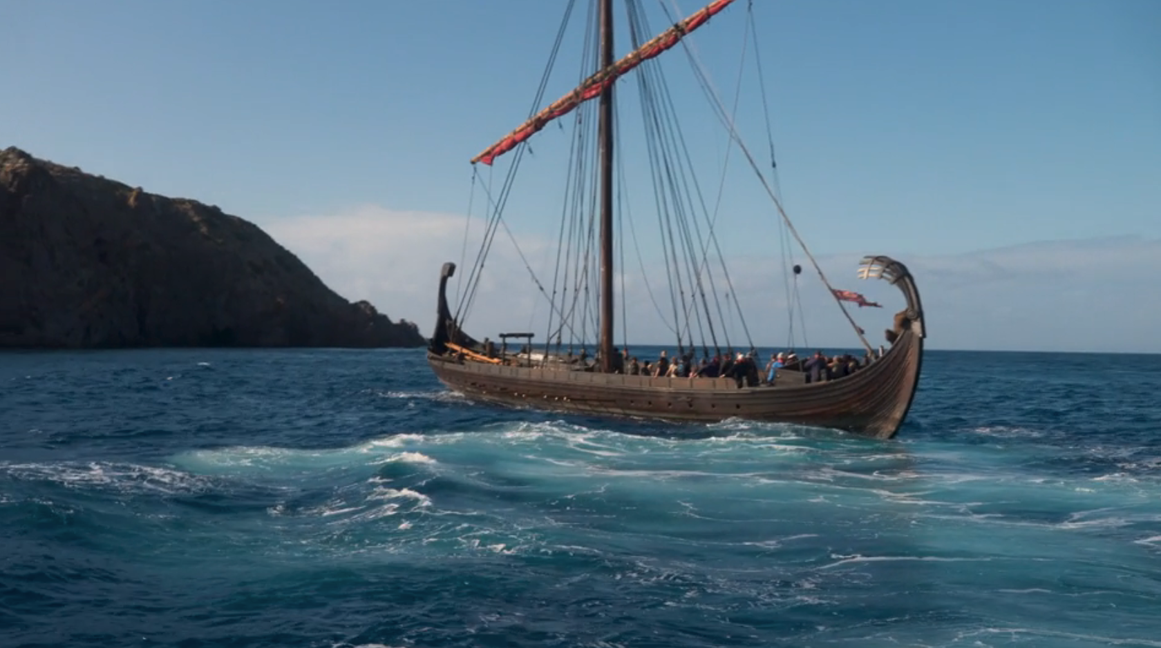 A Viking longship sails on the ocean near rocky cliffs, with several people on board bracing against the wind