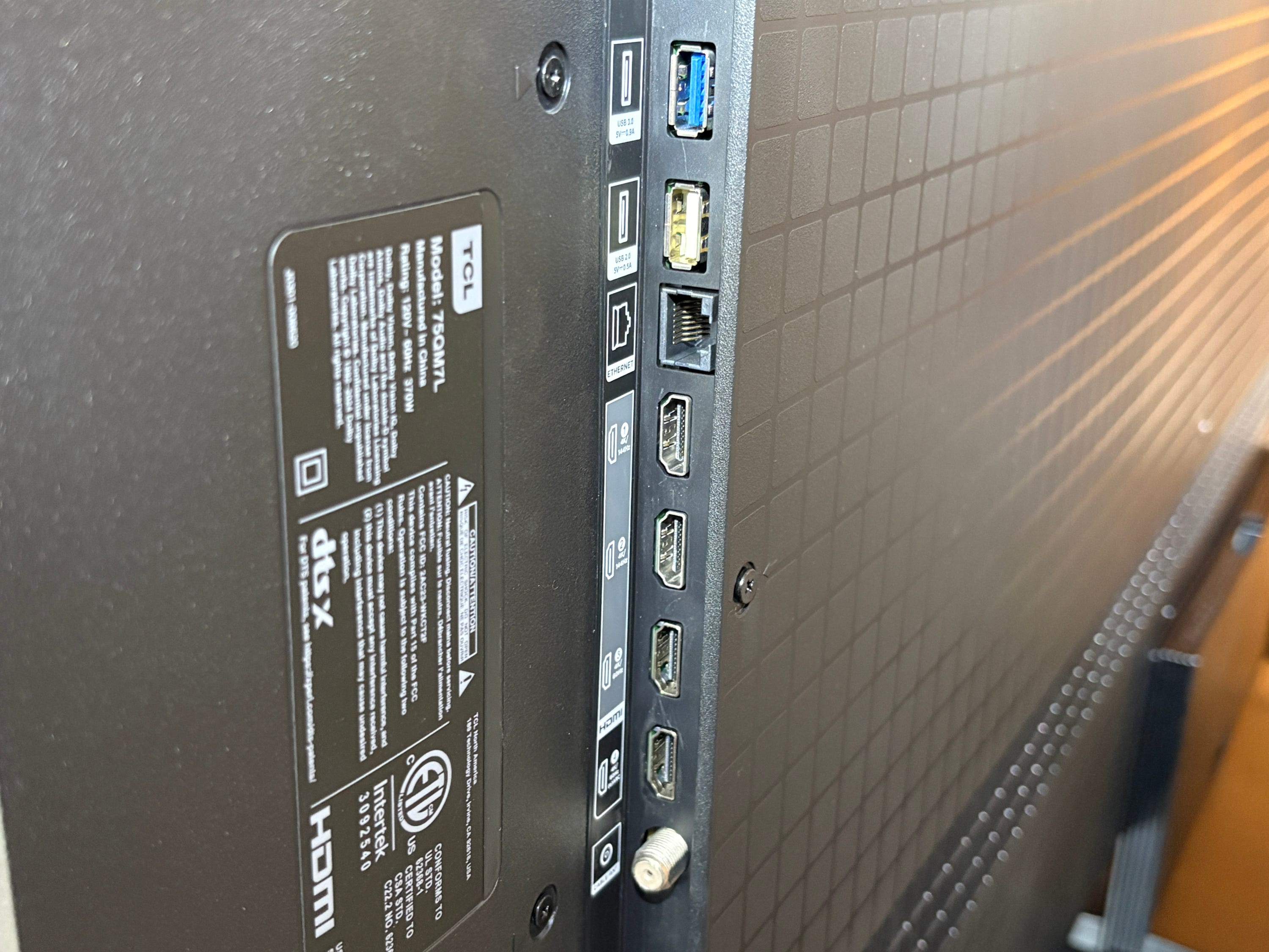 Close-up of the rear of a TCL QM7L TV showing input ports, labels, and a product information sticker.