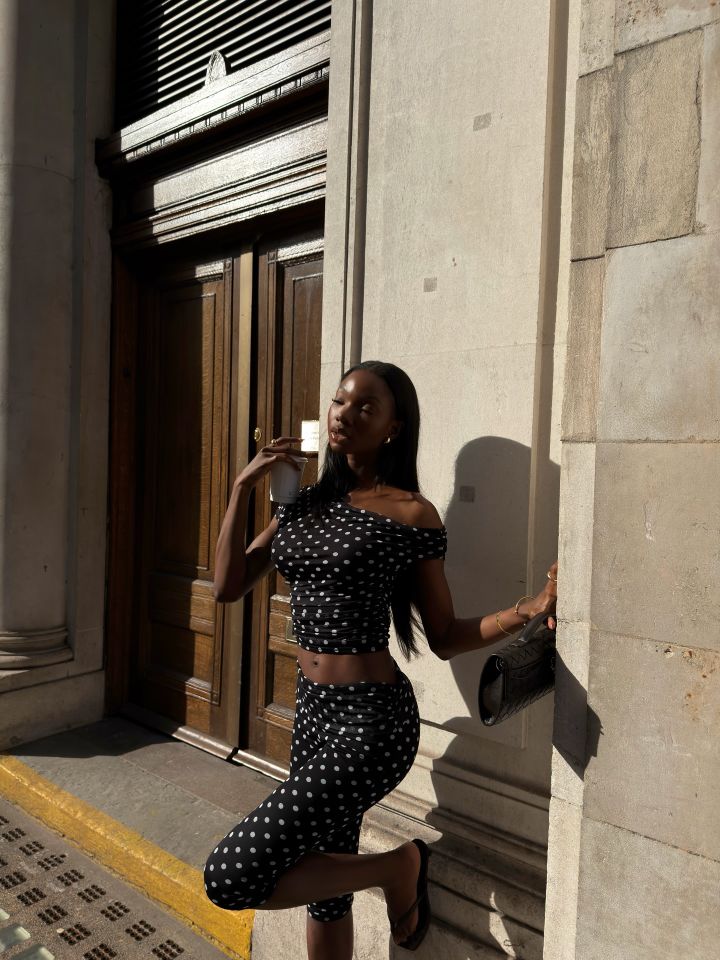 A person poses outdoors against a building wall in a dotted outfit holding a bag