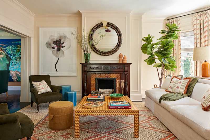 Living room with a white sofa, patterned rug, green plants, and a decorative fireplace with artwork and books on the coffee table.