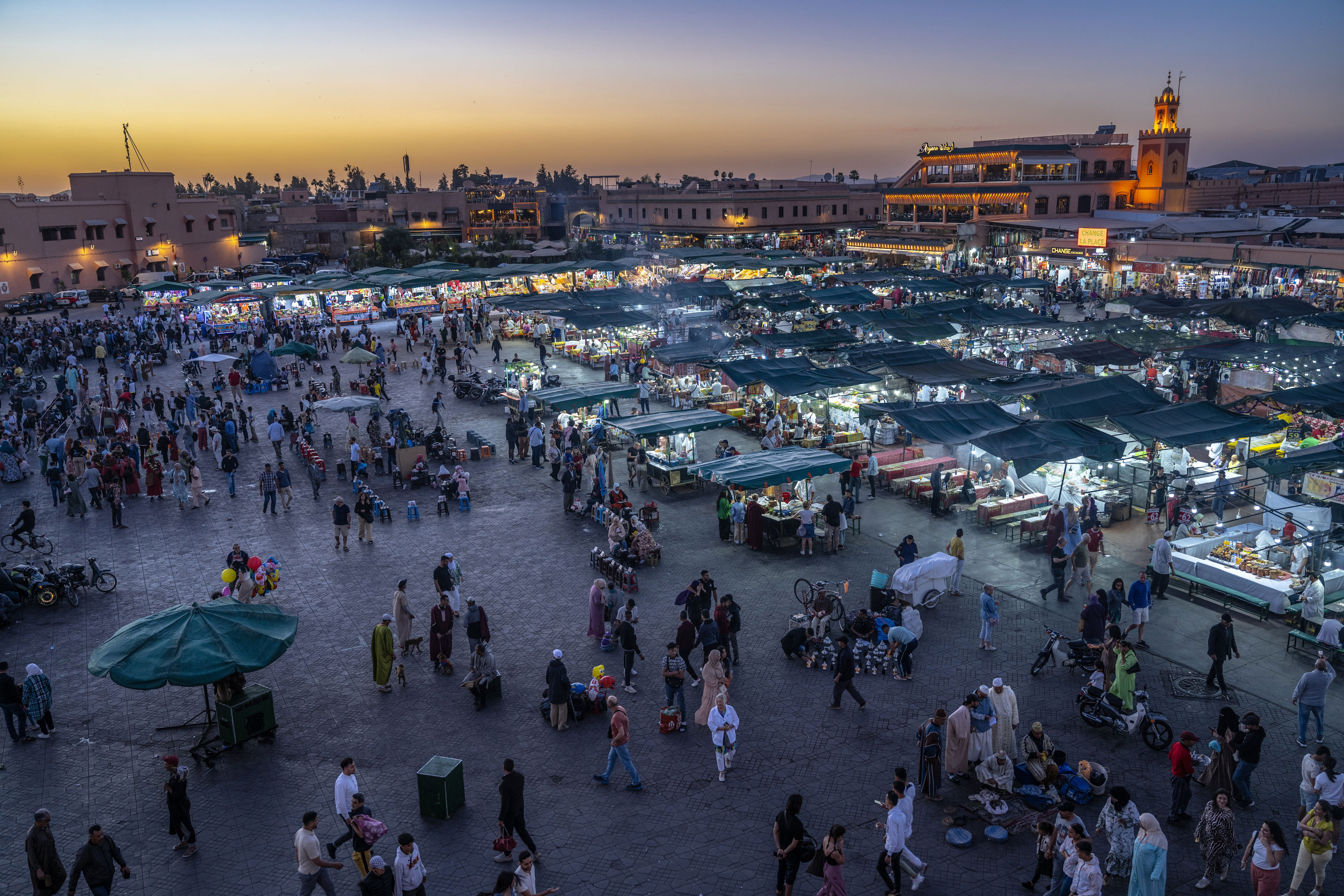 A bustling Moroccan market at dusk, with crowded stalls and people engaging in activities. An illuminated cityscape is visible in the background