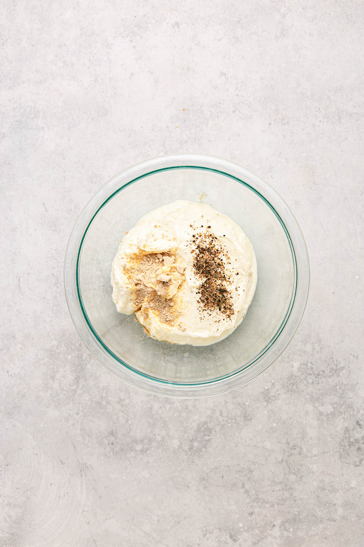 A glass bowl filled with a creamy white mixture topped with ground pepper and a beige seasoning, set on a light gray surface.