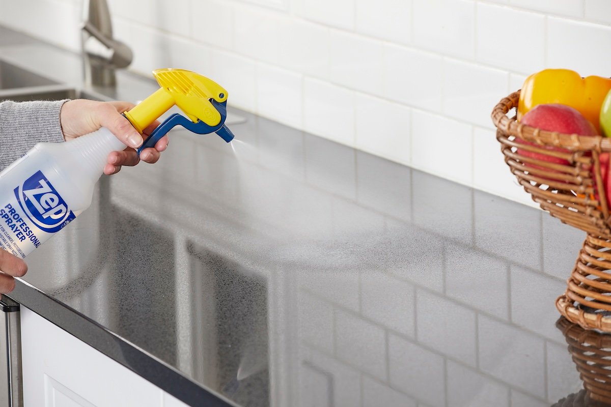 Person sprays grey kitchen countertop with spray bottle near a bowl of fruit.