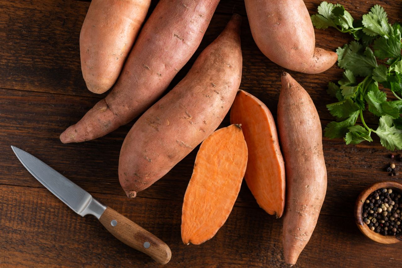 Whole and sliced sweet potatoes on a wooden surface with a knife and garnishes