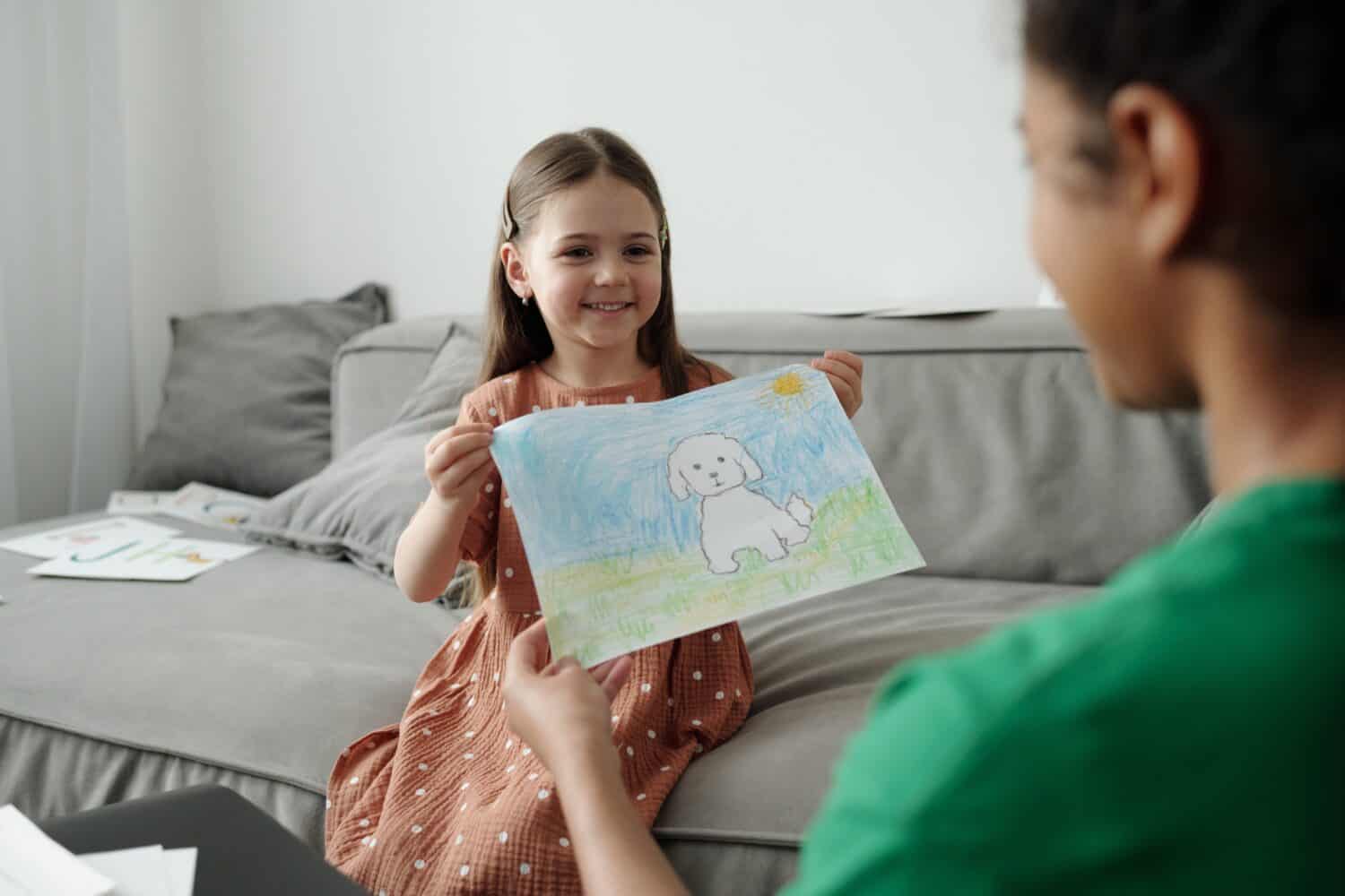 Happy cute little girl sitting on sofa and showing drawing of her pet to African American teenage female sitter while looking at her