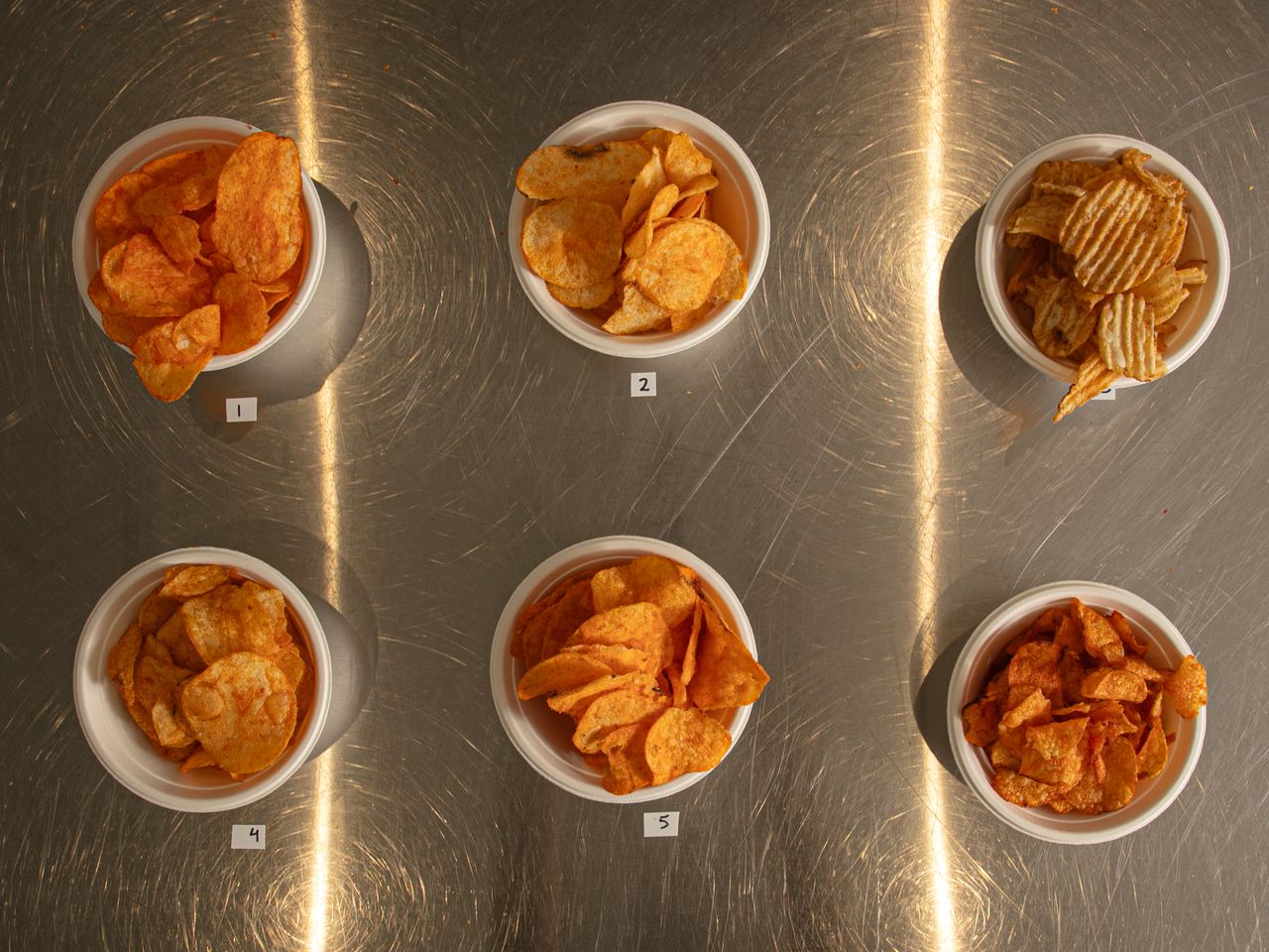 Six bowls containing different varieties of potato chips arranged in two rows on a metallic surface