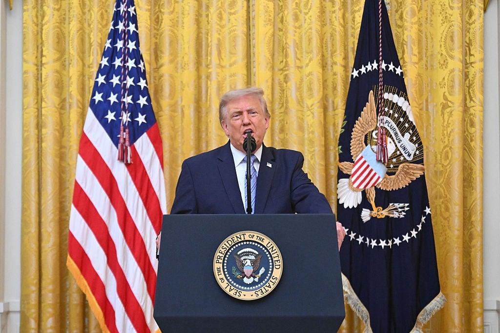 A person at a podium speaks in front of two national flags inside an ornate room, suggesting a formal press event or announcement