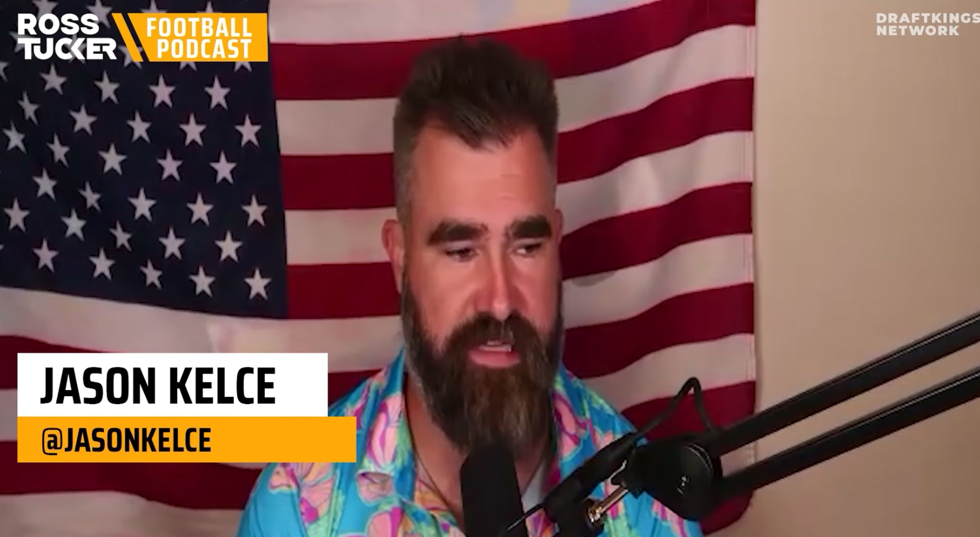 Jason Kelce speaks into a microphone in front of an American flag.