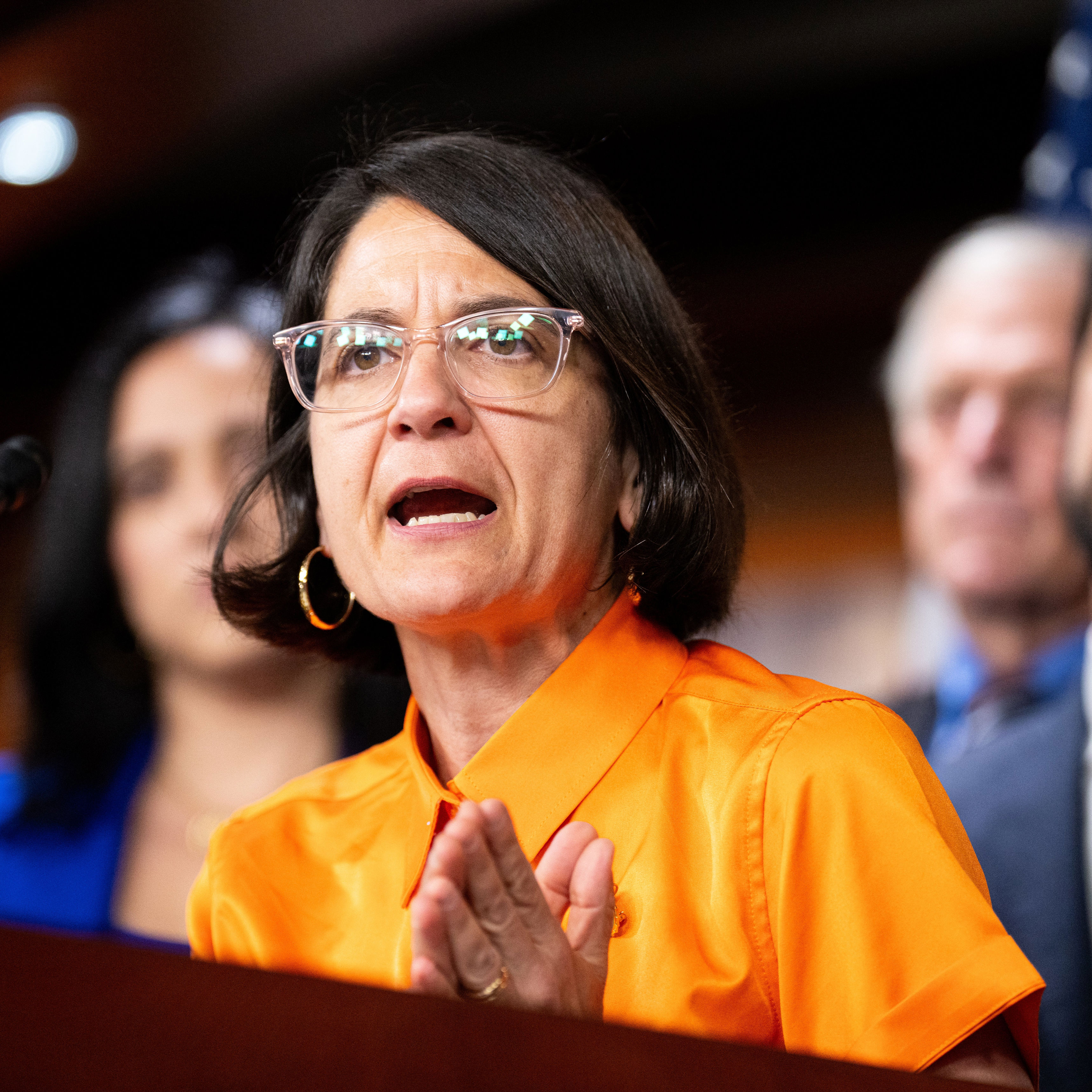 A woman speaks passionately at a podium during a press event, surrounded by others in formal attire