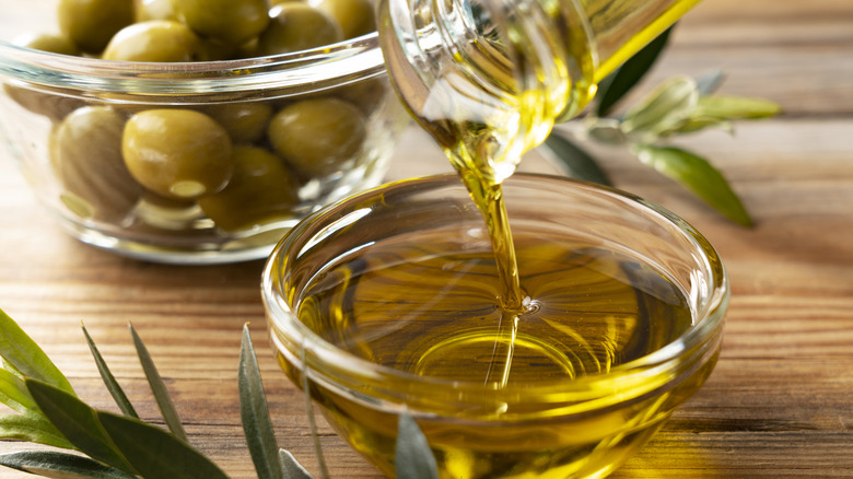 A close-up of olive poured from a glass bottle into a small glass bowl with whole olives in the background