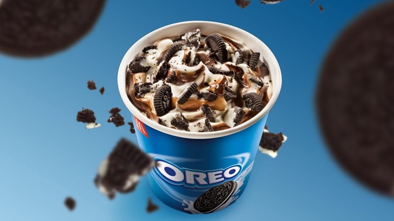 A oreo mcflurry cup against a blue background and oreos hovering all around it