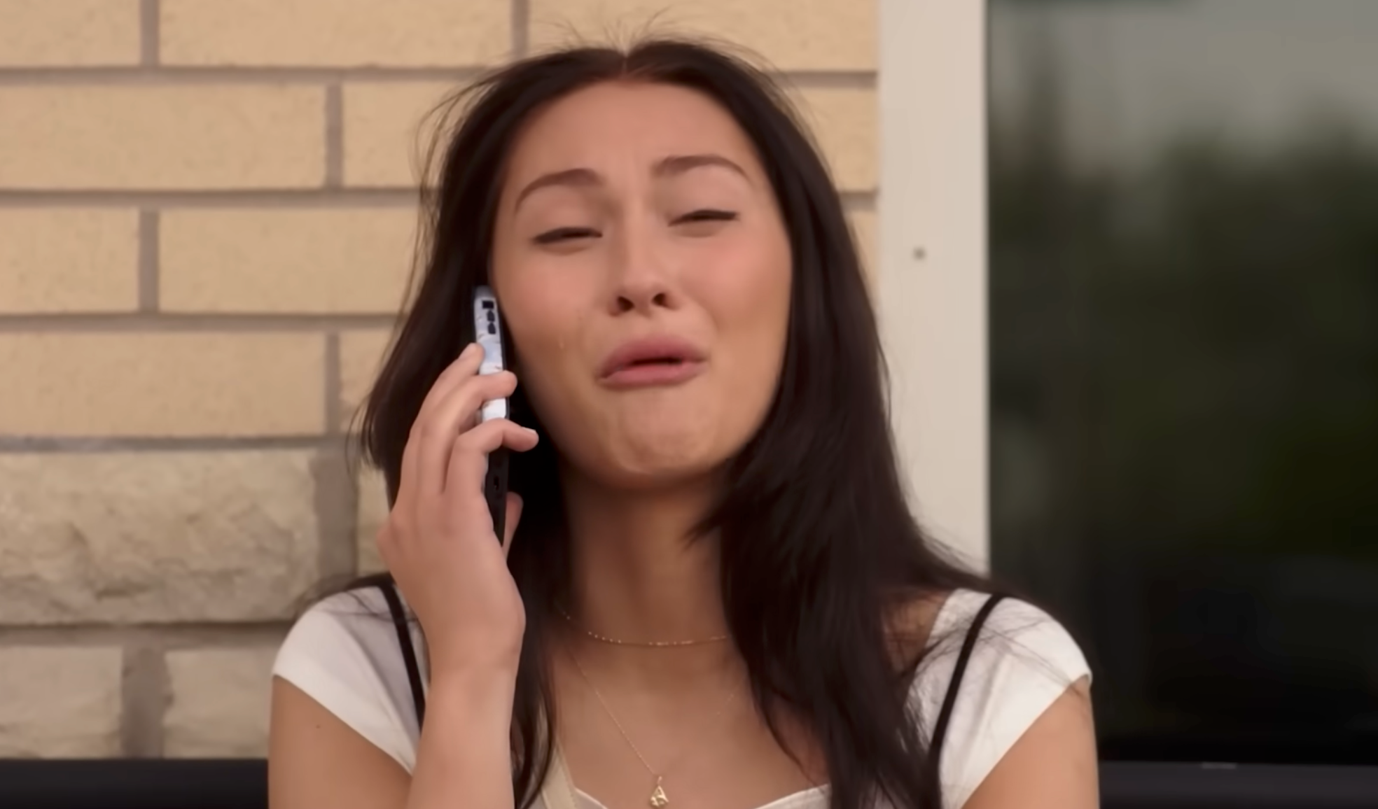 A person sitting outside, speaking on the phone with an emotional expression, possibly laughing or crying