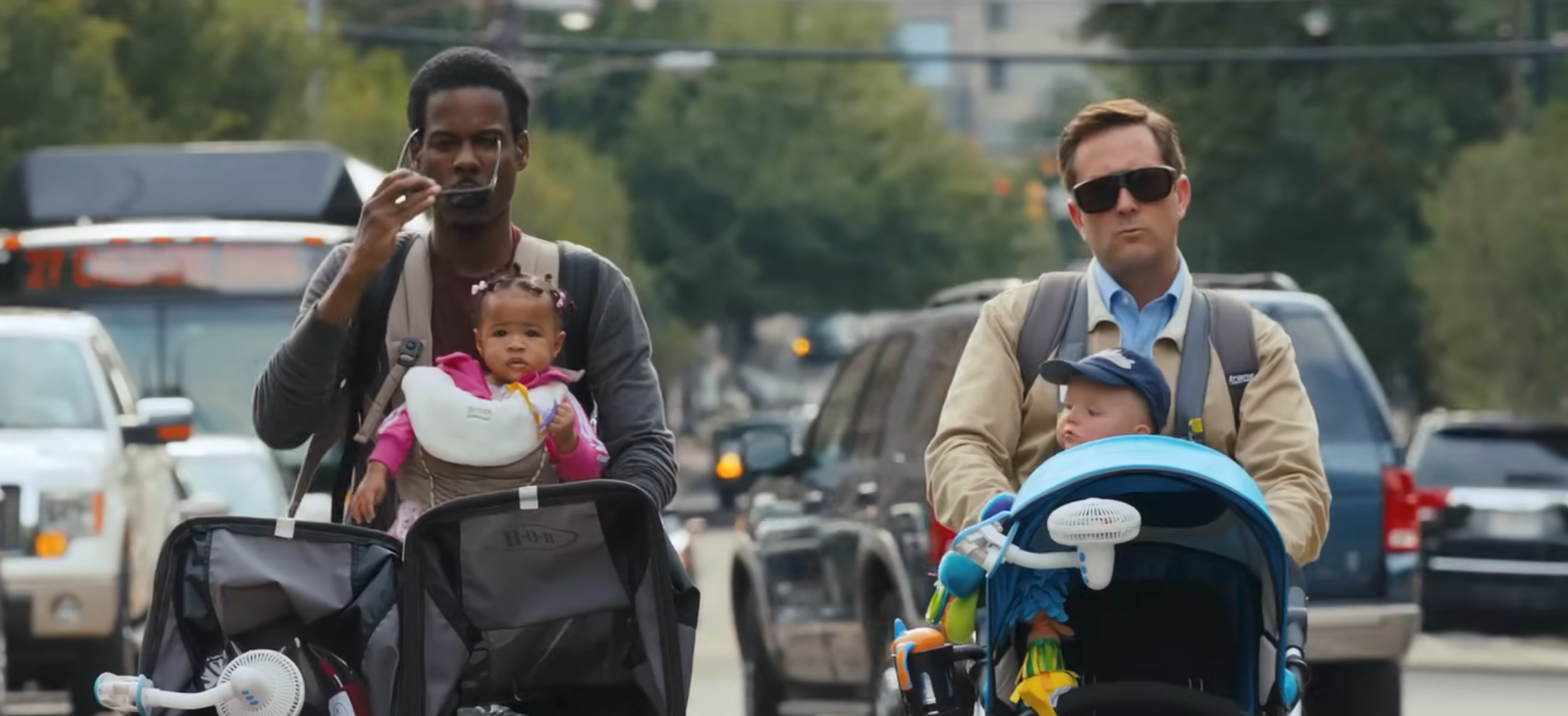 Two men walking with strollers, each carrying a baby. One man wears sunglasses and the other appears engaged in conversation