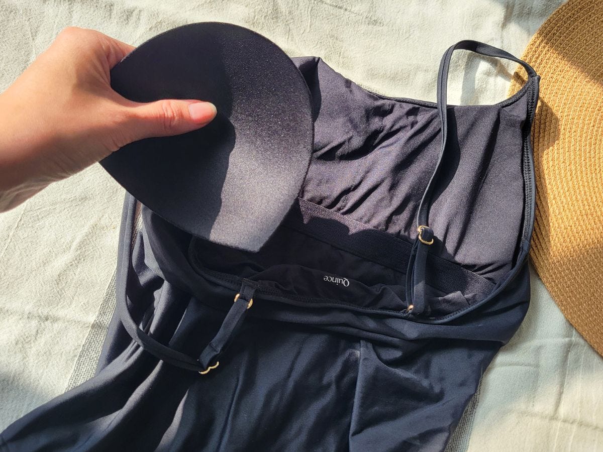 A hand holds a black removable pad above a dark swimsuit beside a straw hat on light fabric.