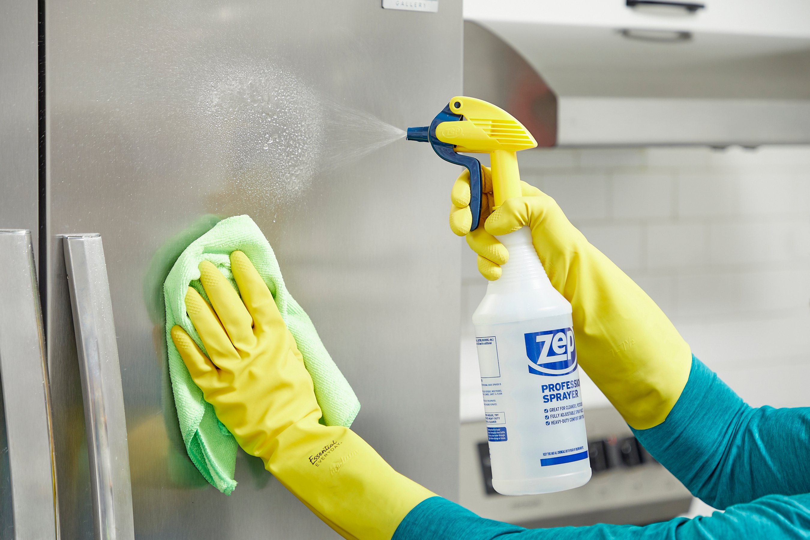 Woman wearing yellow rubber gloves sprays a stainless steel fridge and prepares to wipe it with a microfiber cloth.