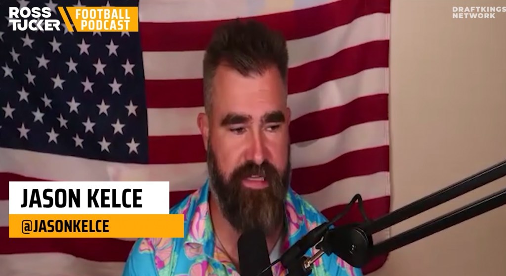 Jason Kelce speaks into a microphone in front of an American flag.
