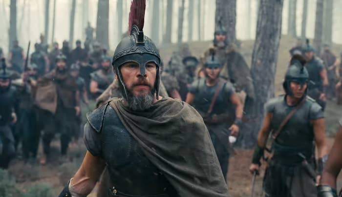 Actors in ancient warrior costumes, with helmets and cloaks, march through a forest setting. The scene evokes a historical battle