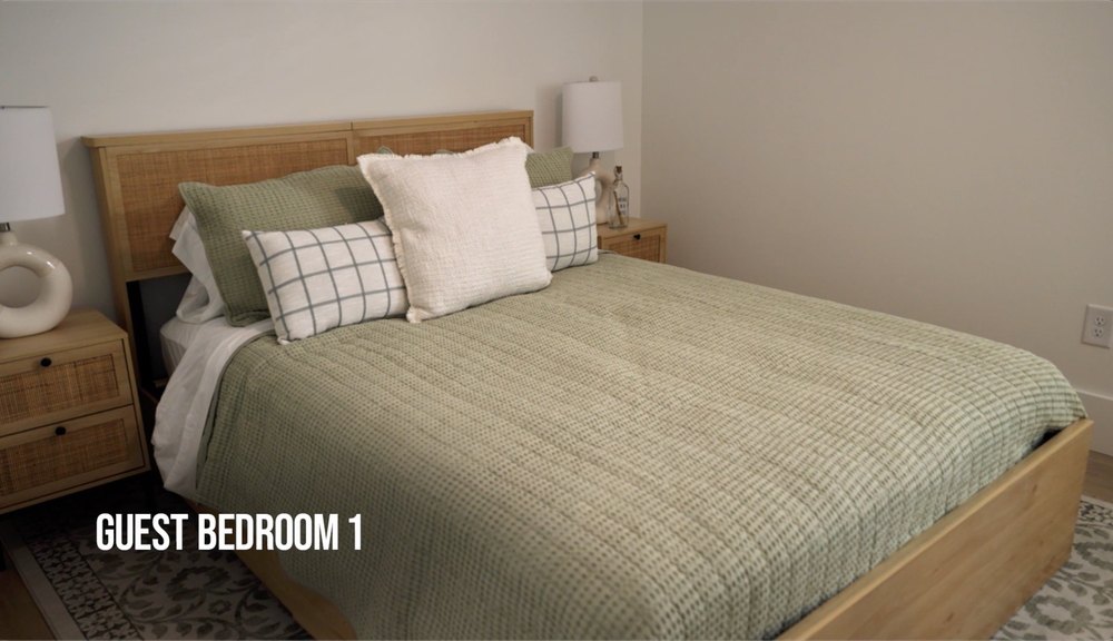 In another bedroom, the home’s palette of neutral finishes continues with creamy walls and sage-colored bedding that covers a wood-toned bed flanked by two wood-toned nightstands.