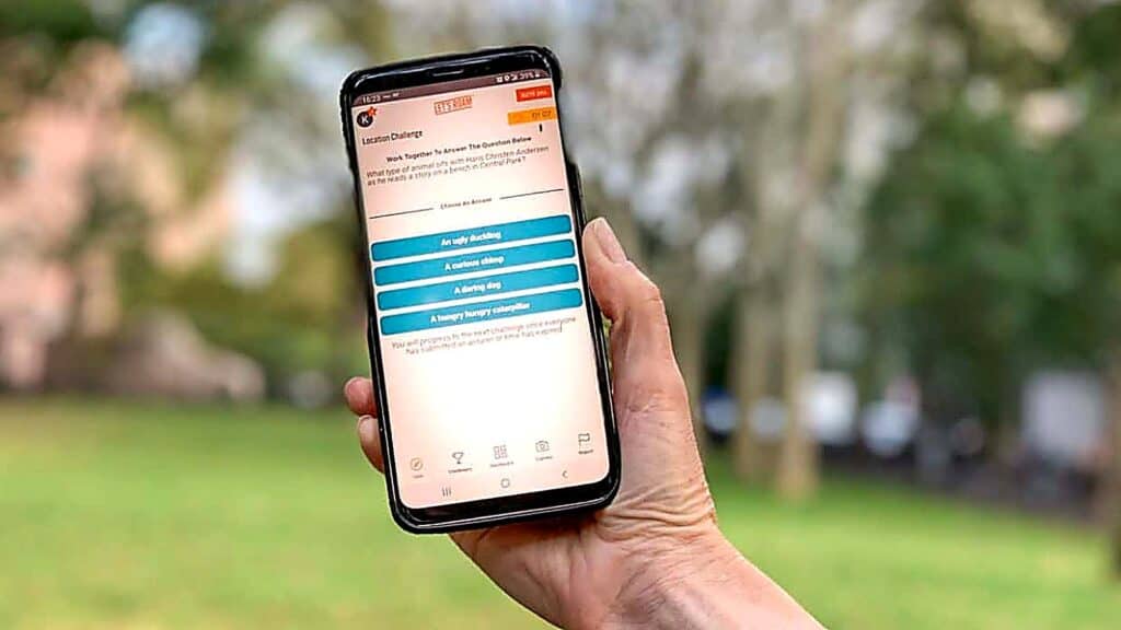 A person holds a smartphone outdoors, displaying a scavenger hunt quiz app with four multiple-choice options on the screen.