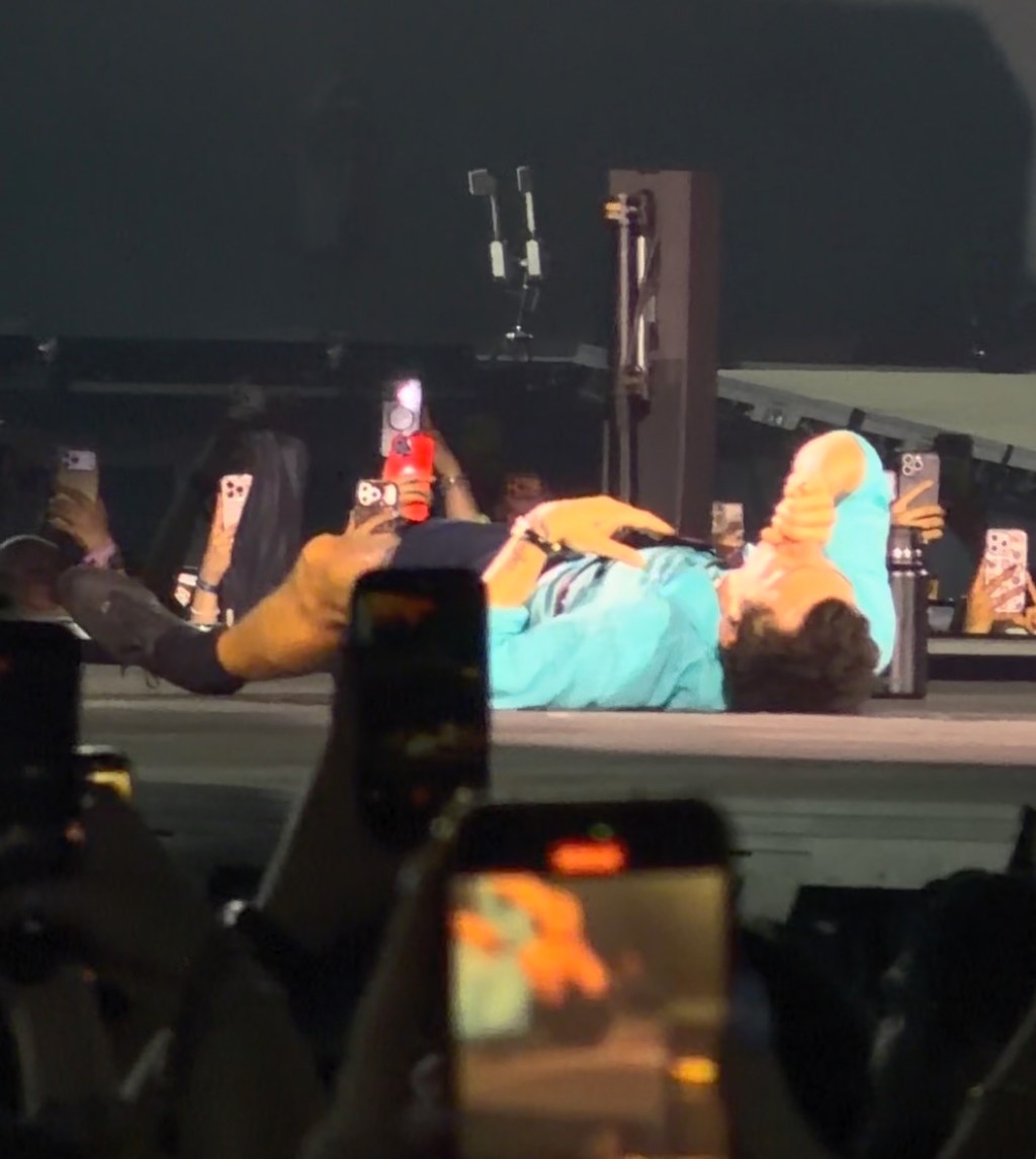 Harry Styles lies on his back on stage, surrounded by fans holding up phones.