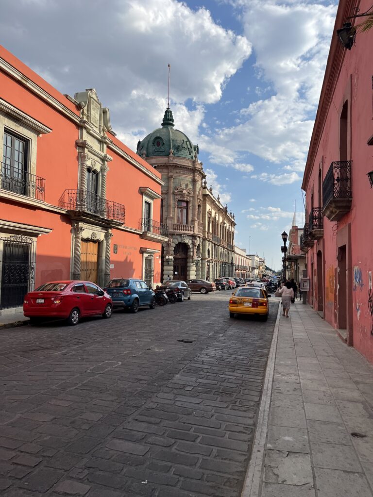 A colorful street in downtown Oaxaca features bright coral and pink colonial buildings framing an ornate historic building topped with a green copper dome. Parked cars, a yellow taxi, and pedestrians bring life to the cobblestone street under scattered afternoon clouds.