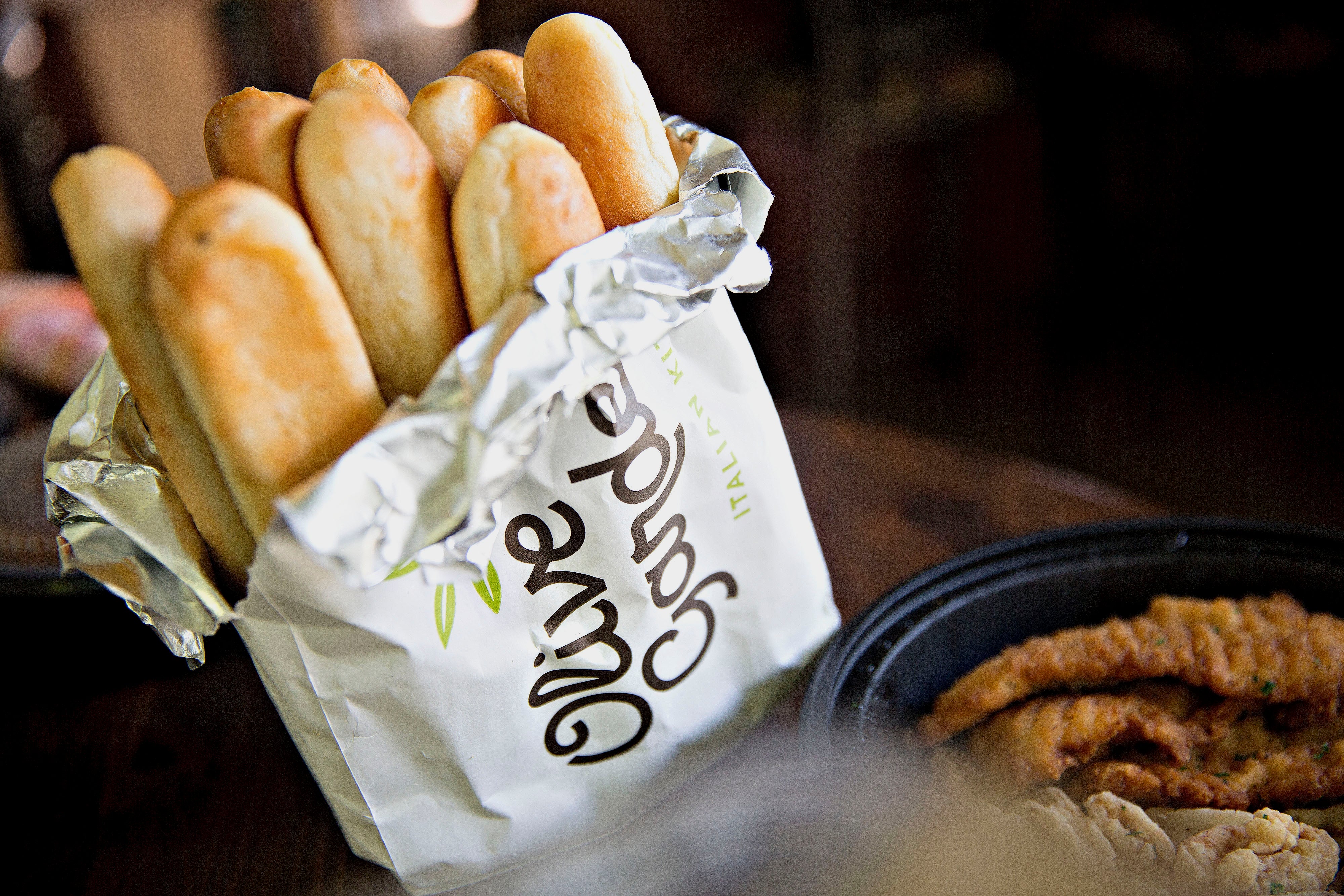 Olive Garden breadsticks wrapped in foil, placed in a branded paper bag, with fried food in a black container nearby