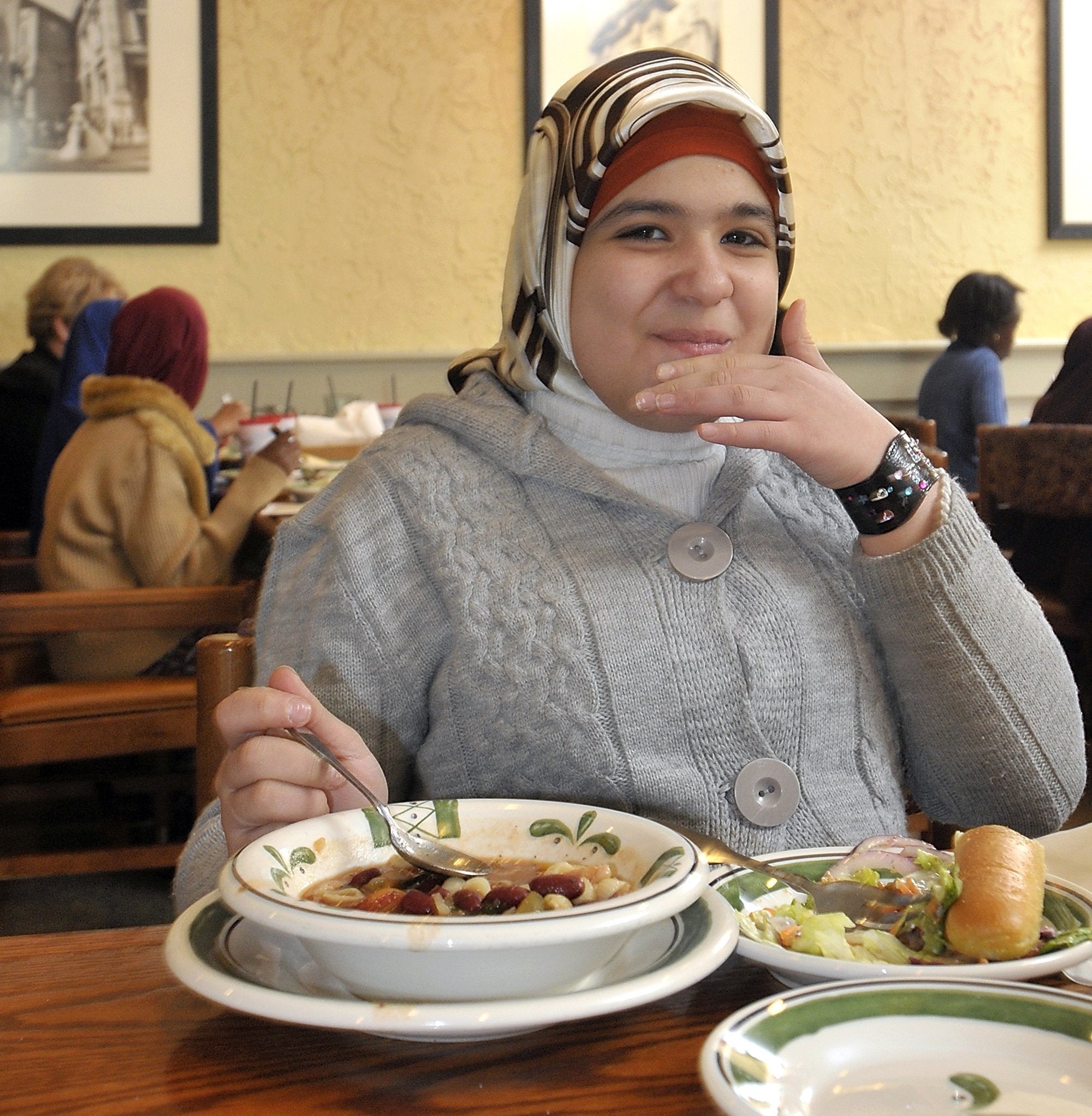 A person sits in a restaurant enjoying a meal, wearing a knitted sweater and a headscarf